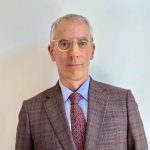 A man with glasses and white hair wearing a brown suit and tie.