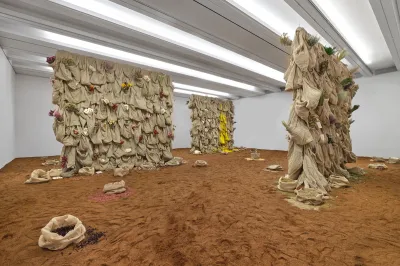 View of a room-size installation featuring sculptures made of hemp bags with flora coming out. The floor is covered in dirt. 