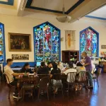 A group is seated at a table in a room with religious stained glass windows.