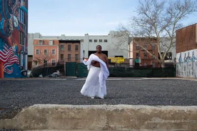 A Black woman with her white top blowing away to reveal her breasts standing in an empty lot.