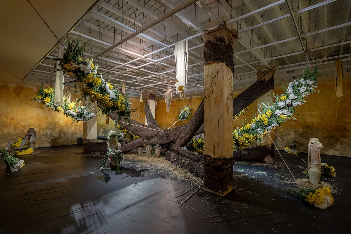 An installation with long wreaths of flowers surrounding columns with fabrics strung around them.