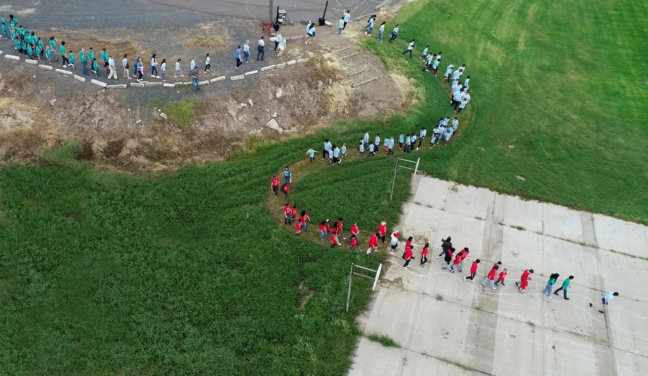 Aerial drone shot of school children of varying ages walk in pairs in a curving line in the shape of a river. The students wear matching t-shirts in turquoise, sky blue, or red that bear the project