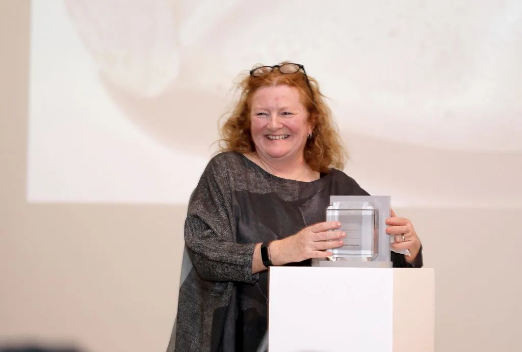 A woman with shoulder-length wavy red hair and glasses on her head, weaing a black dress, smiling while standing behind a plinth and holding a glass award