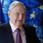 A man in a suit and tie in front of a blue flag.