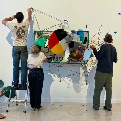 Three people install a wall-hung sculpture on a wall. 