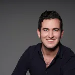 A headshot of ARTnews senior editor Maximilíano Durón. He is smiling and wearing a black shirt.