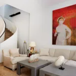 A modern looking living room with white sofa and grey tables. On the wall is a big red drippy painting of a nurse in a face mask.