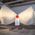 A sculpture of a giant white bra with a red flower in the center has a tag that reads: ya ho nay pecho que aguante.