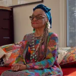 A Black woman in a blue turban and a multicolored shirt seated on a couch with patterned cushions.