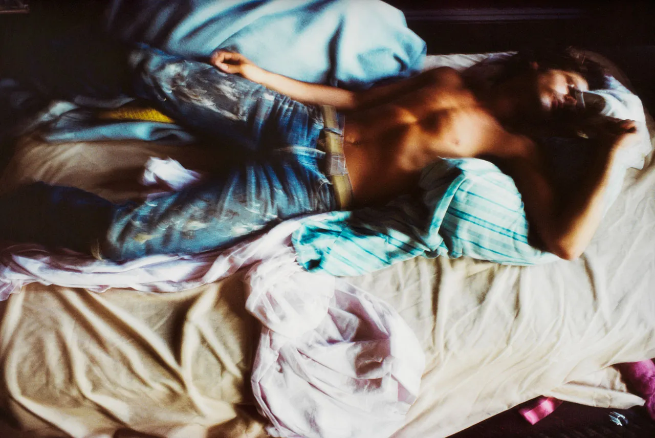 A still image from a video showing a shirtless man in jeans laying on an unmade bed.