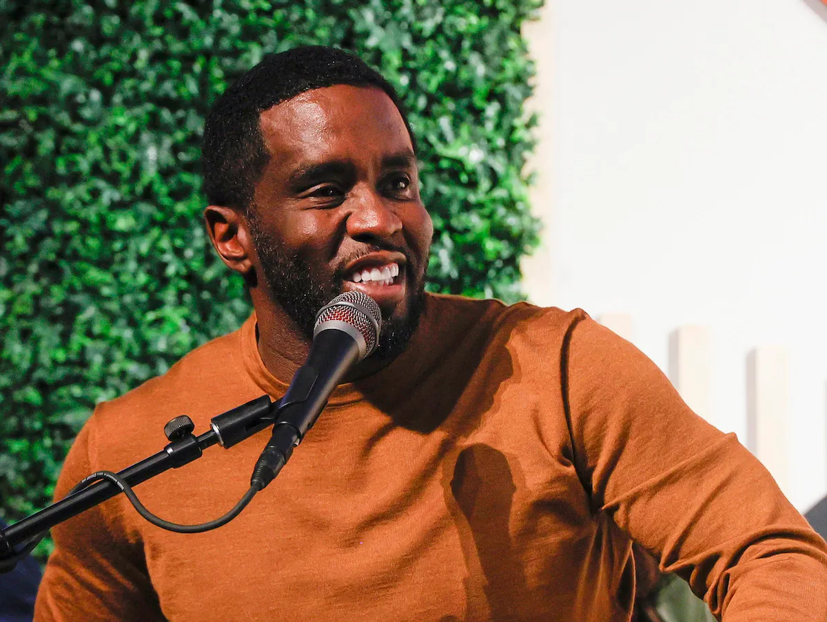 A man in a brown sweater speaking into a microphone.