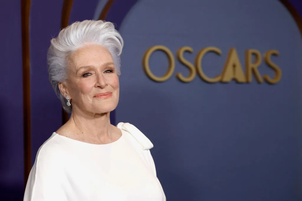 Close-up photo of a woman with white hair and a while dress, standing in front of a dark blue wall with the word