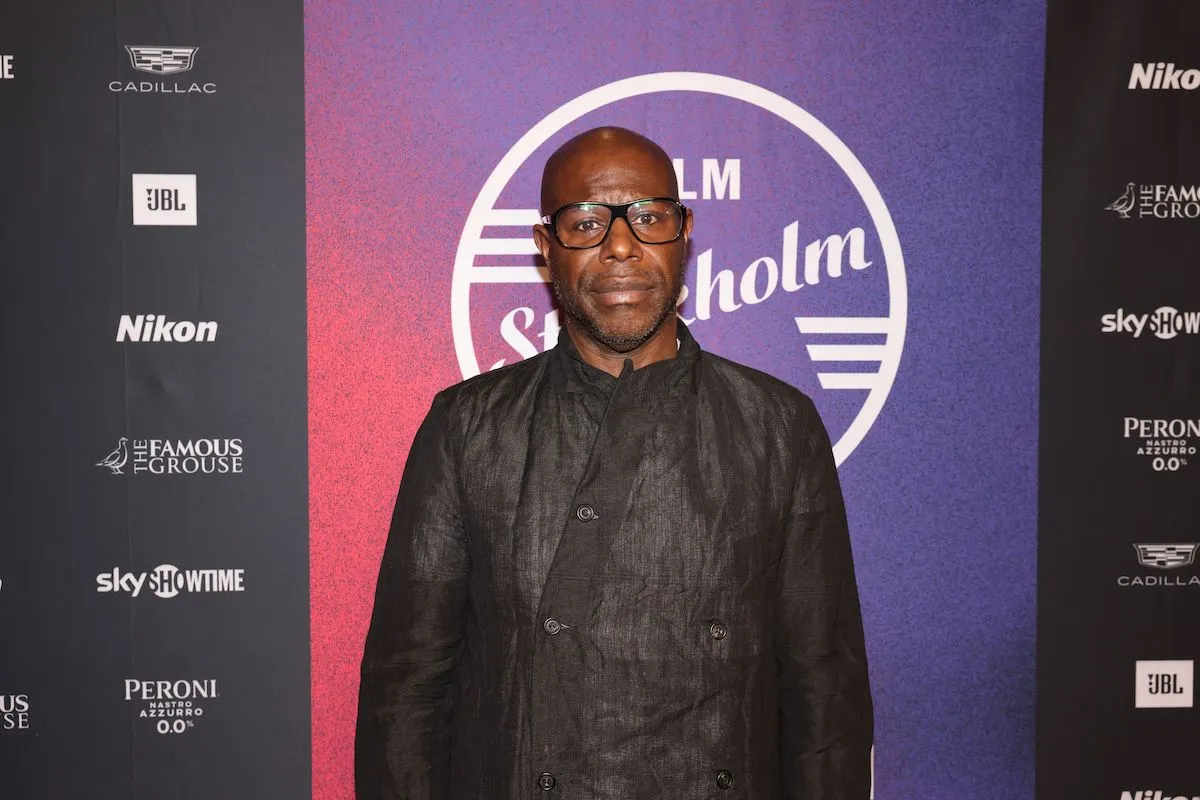 Portrait of Steve McQueen, a Black man in a black suit standing on a red carpet.