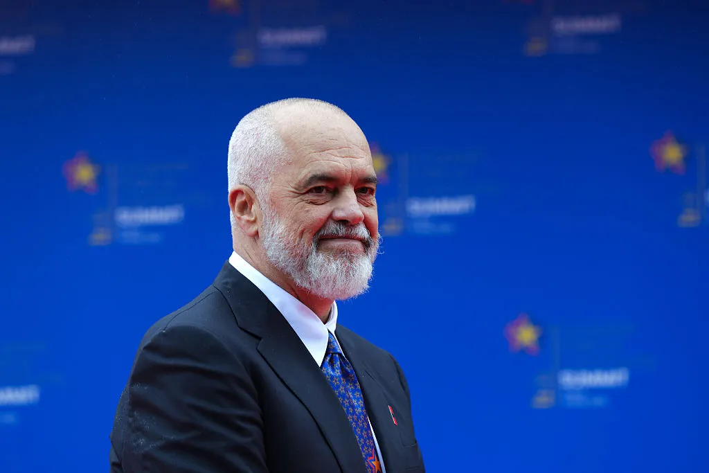 A bald man with a cropped white beard and buzzed hair in a dark suit and blue tie, standing in front of a blue background and smiling
