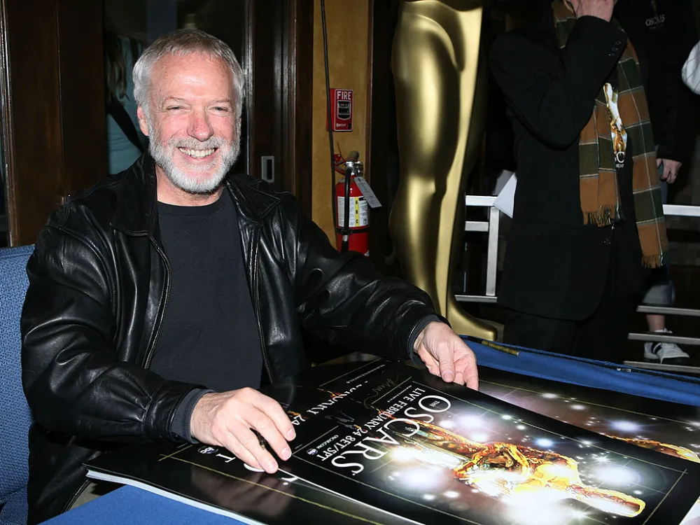 A man with white hair and a close-cropped white beard smiling, sitting at a table with a poster of a gold award on the table in front of him