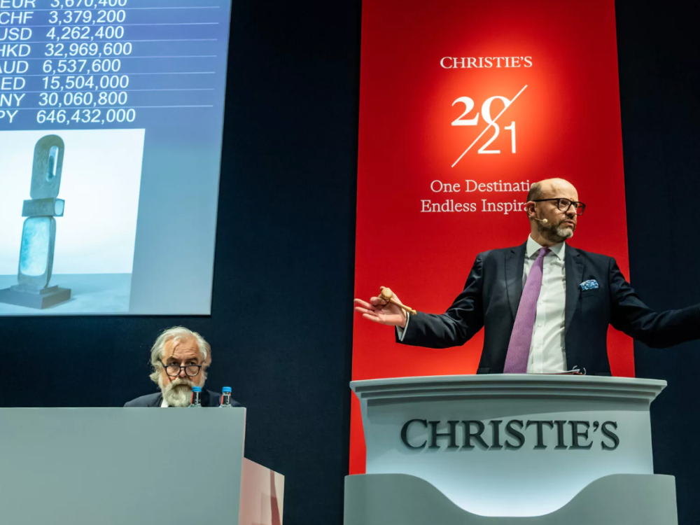 Two men sell art from behind rostrums at an auction.