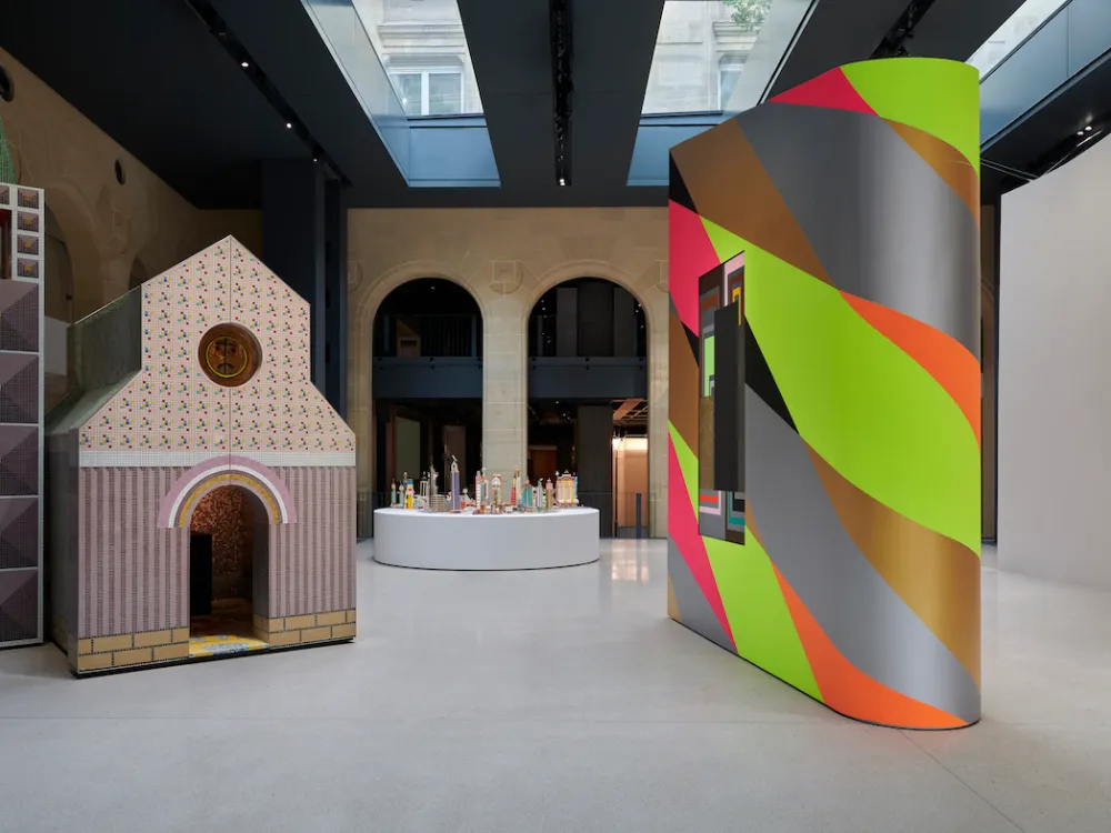 A gallery with a tall house-like structure and a freestanding wall painted with a shimmery green and red pattern.