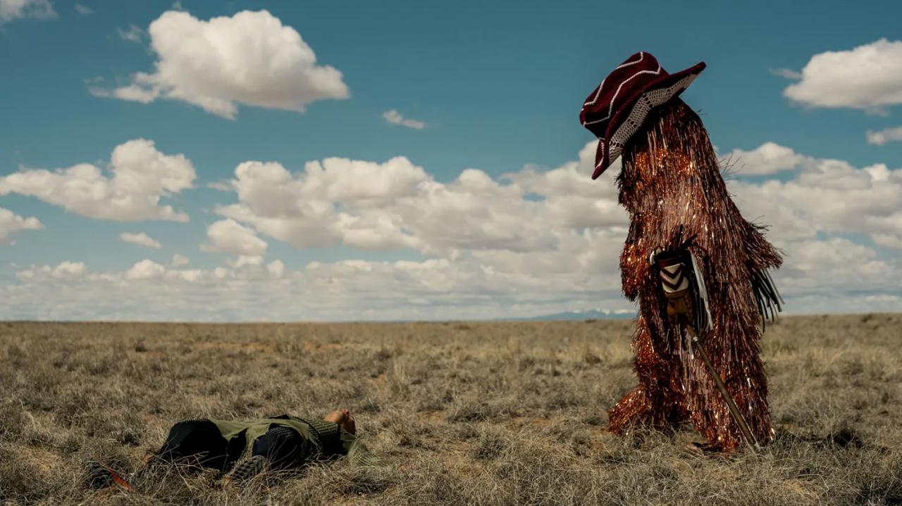 A mascot covered in tinsel and wearing an oversize cowboy hat walks across a desert.