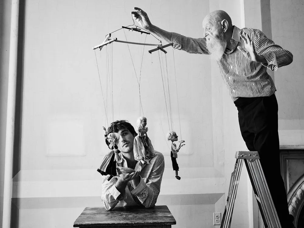 An older man stands on a step stool holding three marionette figures over a younger man who looks at them.