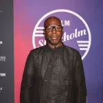Portrait of Steve McQueen, a Black man in a black suit standing on a red carpet.