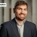 A man with a dark grey blazer and grey shirt smiles at the camera.