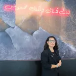 A woman with long dark hair, glasses, and a black suit stands in front of a blue and gray patterned wall with Arabic text in red neon above her head