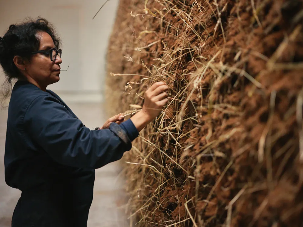 Delcy Morelos, wearing a blue jumpsuit, places hay into a large sculpture made of earth.