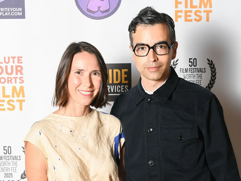 A man and woman standing next to each other smiling. The woman has chin-length brown hair and a cream-colored short-sleeve dress with blue lines; the man has short dark hair, dark glasses, and a black top and pants.