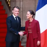 A man in a dark blue suit and tie smiling and shaking hands with a smiling woman in a red dress and jacket, standing in front of a red staircase, with flags in the background