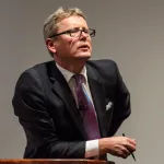 A man in a suit leaning forward on a rostrum. He holds a pen in one hand.
