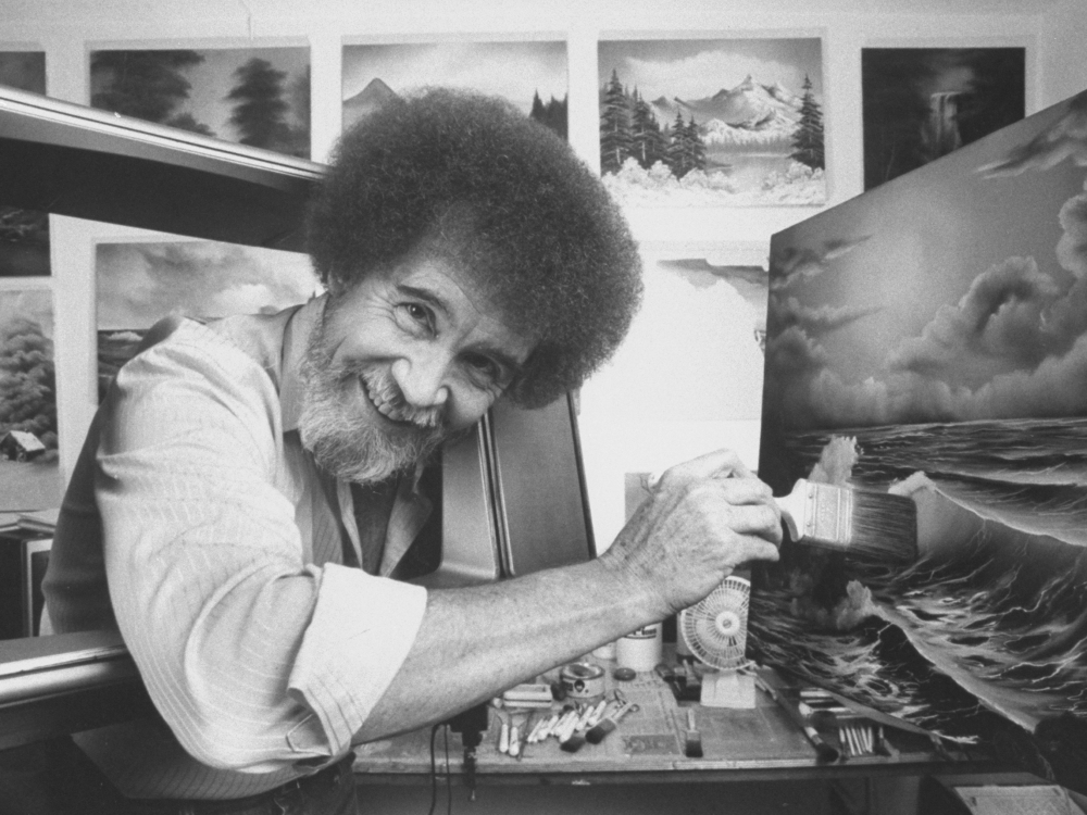 black and white photo of a man with bushy hair looking at the camera and holding a paintbrush, leaning towards a canvas