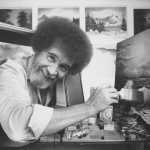 black and white photo of a man with bushy hair looking at the camera and holding a paintbrush, leaning towards a canvas