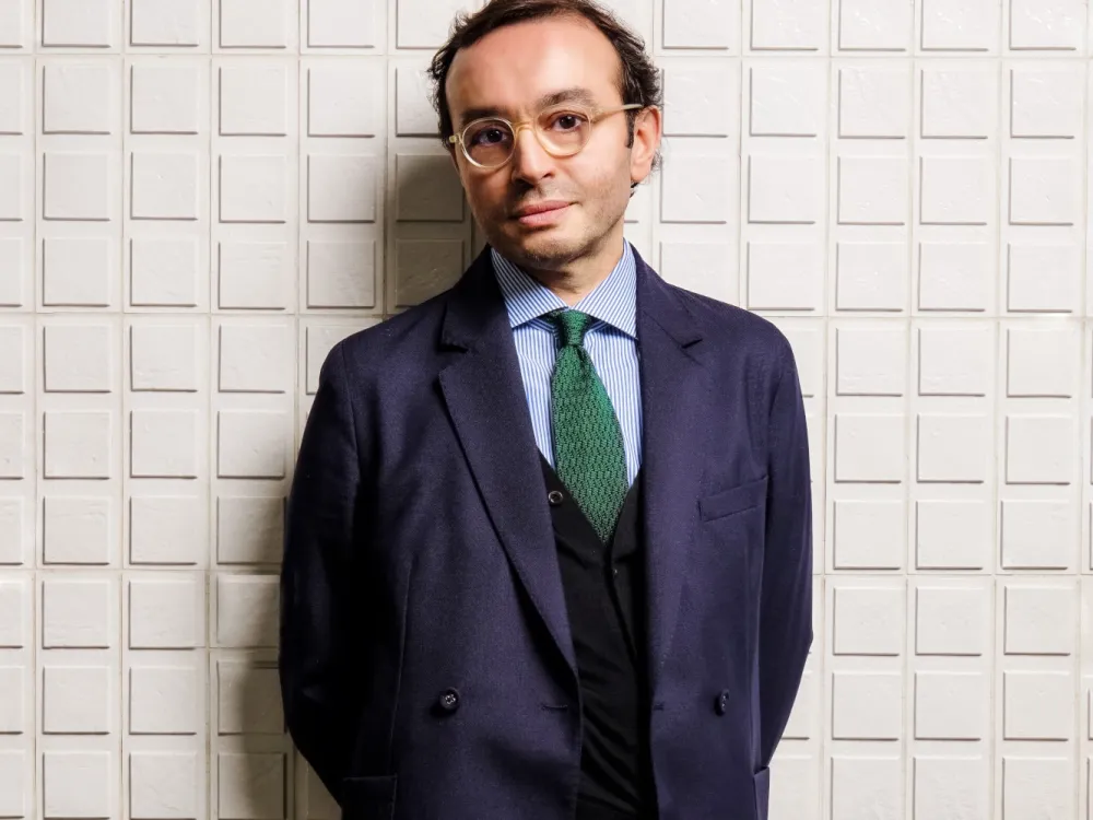 A photo shows a man, art dealer Mehdi Chouakri, posing for the camera in a blue suit, black vest, and green tie, wearing stylish glasses.