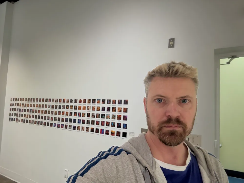 A young white man, artist Nick Dwyer, poses in an art gallery with a number of small images pinned to the wall behind him