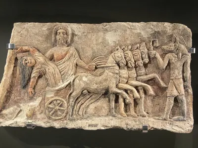 A limestone relief showing a woman being dragged by a man in a chariot pulled by four horses with Anubis at right.