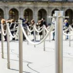 Stainless steel poles linked by grey ropes for queue control at the entrance of a tourist site, with drop shadows on a light ground and blurry people queueing up in the background.