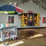 View of a university gallery exhibition with paintings on the wall and a sculpture in the foreground.