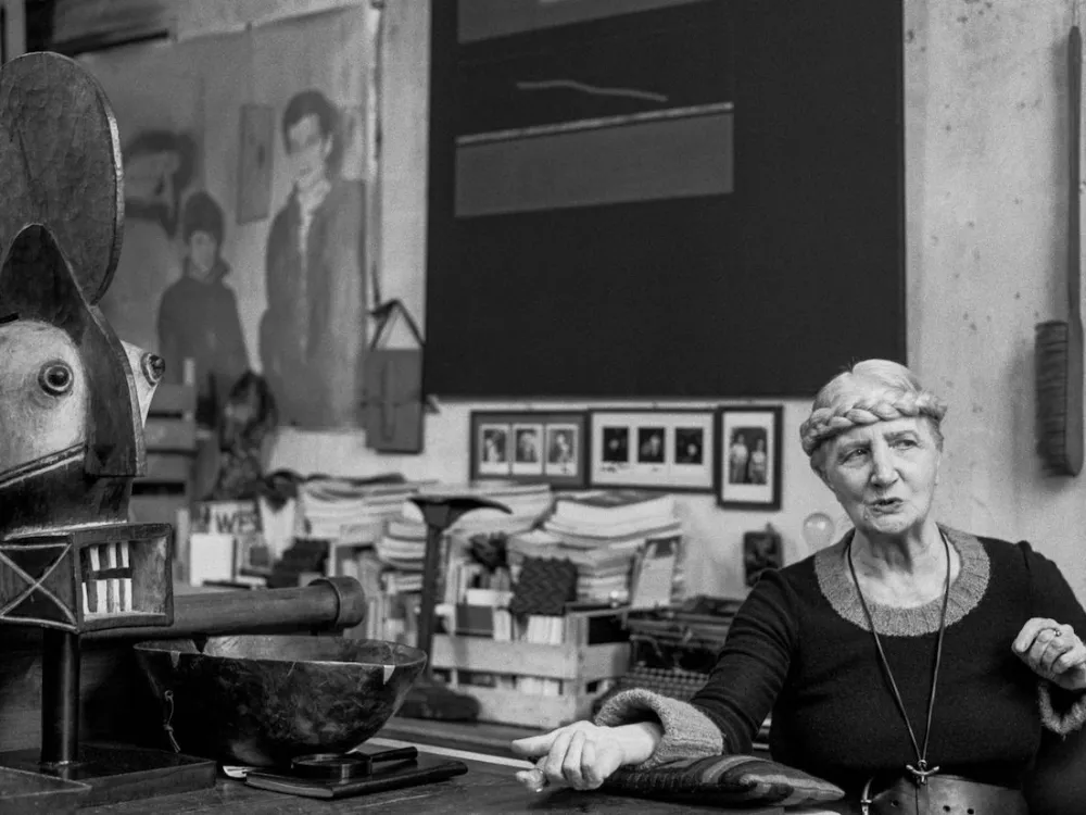 A woman with her braided blond hair tied around her head seated in a studio.