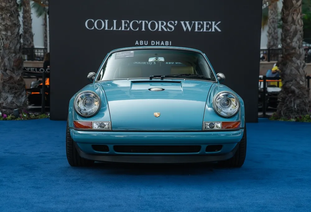 A light blue Porsche on a dark blue carpet surrounded by trees in the background.