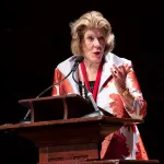 A woman in a dress speaking at podium with a microphone.