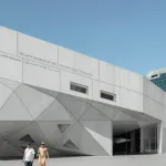 exterior of an angular gray modernist building with geometric panels and two figures walking in the foreground