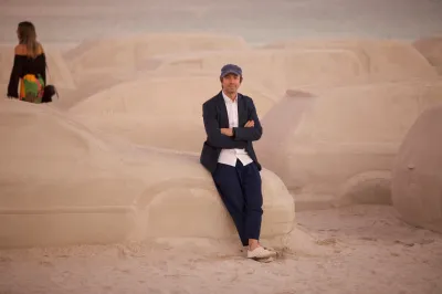 An artist, Leandro Erlich, wears a black suit and baseball cap as he poses with multiple sand sculptures of cars and trucks.