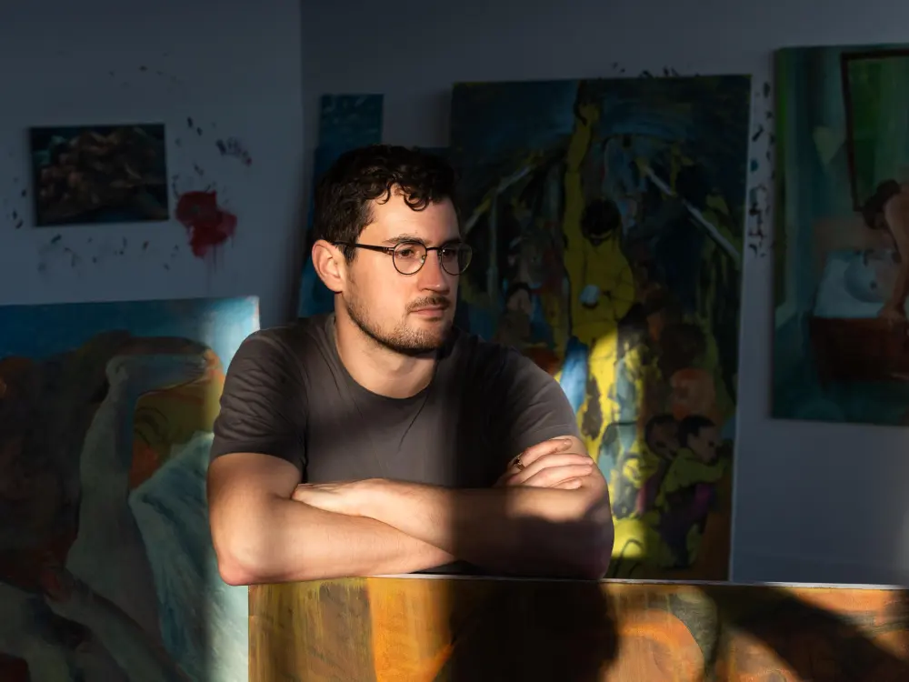 Portrait of Louis Fratino with his arms crossed atop a painting cast mostly in shadow.
