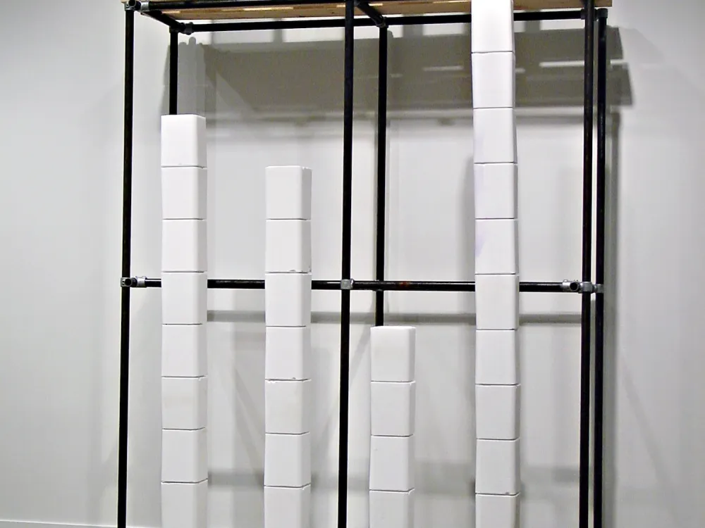 A sculpture against a white wall consists of a metal frame containing several stacks of white cubes.