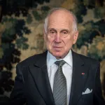 A man in a suit and tie before a large tapestry of a forest.