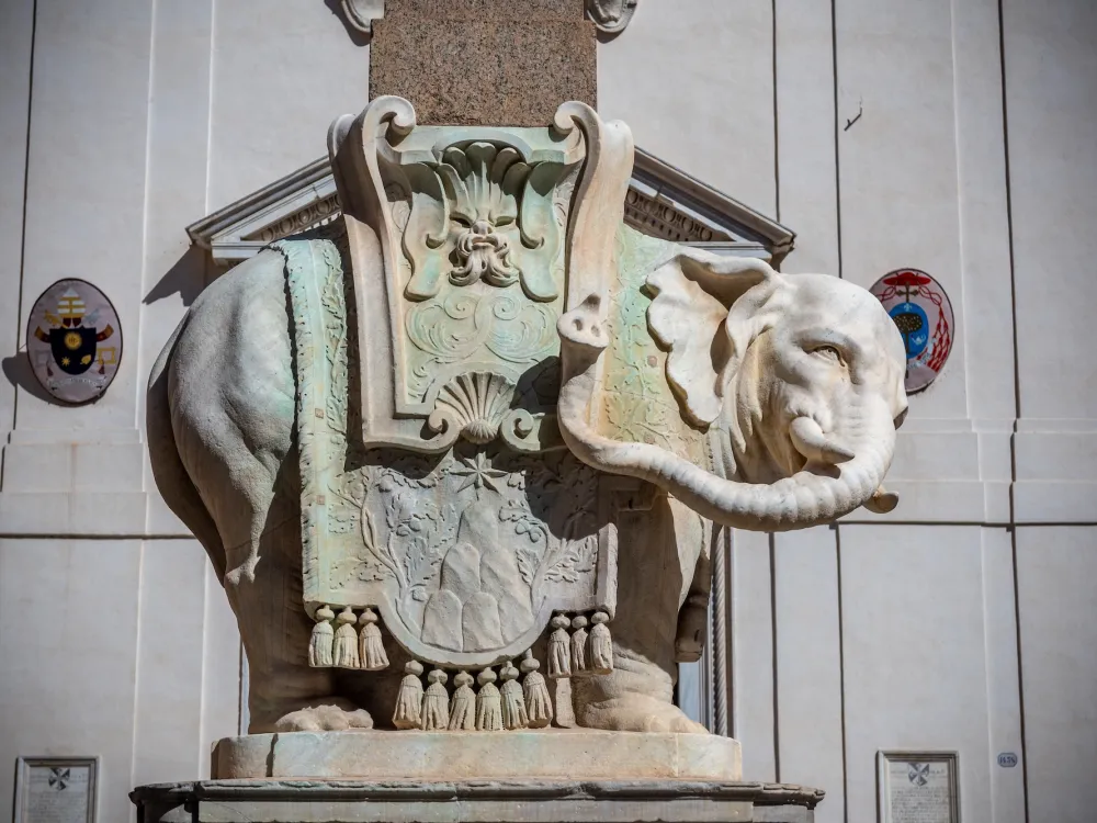 A photo shows a marble sculpture of an elephant in elaborate dress standing in a piazza in Rome, in front of a marble wall