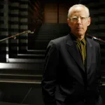 A white man, middle-aged, with receding gray hair, stands in a dark setting wearing a suit and clear-framed glasses.