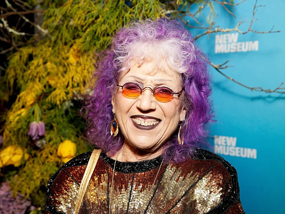 The artist Judy Chicago, sporting purple hair and sunglasses, smiles for the camera in front of a wall printed with the name of the New Museum