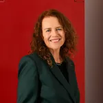 a smiling woman with wavy shoulder-length reddish hair, wearing a dark green blazer and standing in front of a red wall