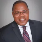 A Black man in a suit and glasses smiles at the camera.